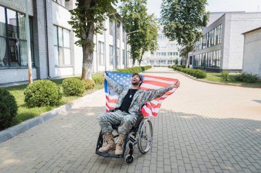 overjoyed handicapped military man in wheelchair holding usa flag and laughing on city street clipart