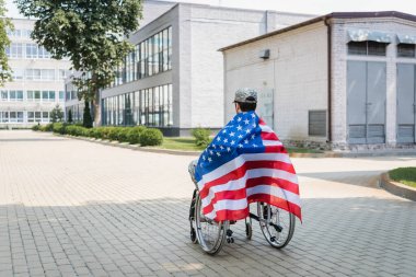 back view of young veteran, covered with usa flag, sitting in wheelchair in city clipart