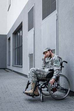 disabled military man in wheelchair holding bottle of whiskey near grey building outdoors clipart