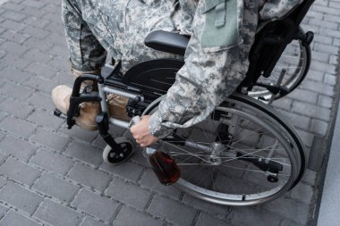 partial view of handicapped military man holding bottle of alcohol while sitting in wheelchair outdoors clipart