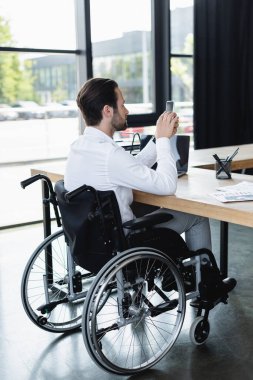 Tekerlekli sandalyedeki engelli işadamı ofisteki dizüstü bilgisayarın yanında akıllı telefondan mesajlaşıyor.