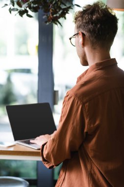 Gözlüklü ve kulaklıklı serbest çalışan. Kafede boş ekranlı dizüstü bilgisayar kullanıyor.