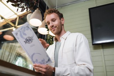 low angle view of cheerful young man looking at blurred menu in cafe clipart