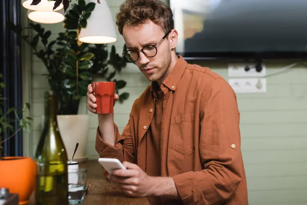 Joven Gafas Sosteniendo Taza Mensajes Texto Teléfono Inteligente Cafetería —  Fotos de Stock