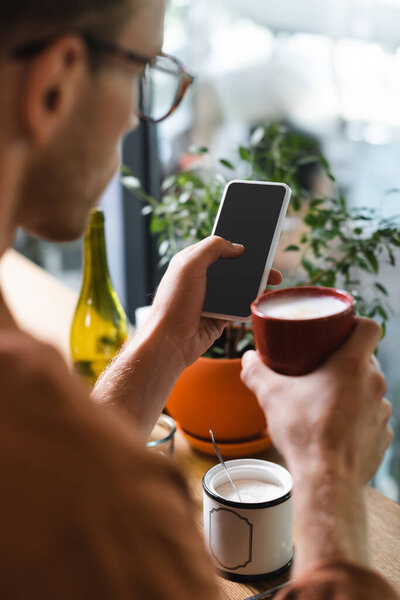 blurred young man in glasses holding cup of coffee and texting on smartphone in cafe 