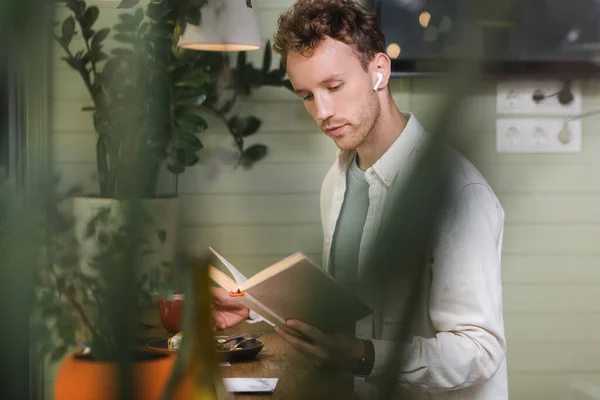 Krullende Man Draadloze Oortelefoons Lezen Boek Wazig Cafe — Stockfoto