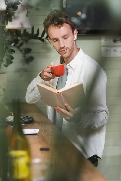 Curly Man Wireless Earphones Reading Book While Holding Cup Blurred — Stock Photo, Image