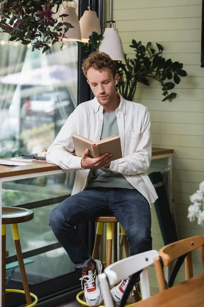 Krullende Man Draadloze Oortelefoons Lezen Boek Buurt Menu Tafel Cafe — Stockfoto