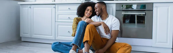 若いアフリカ系アメリカ人のカップルが笑顔でキッチンの床に座って バナー — ストック写真