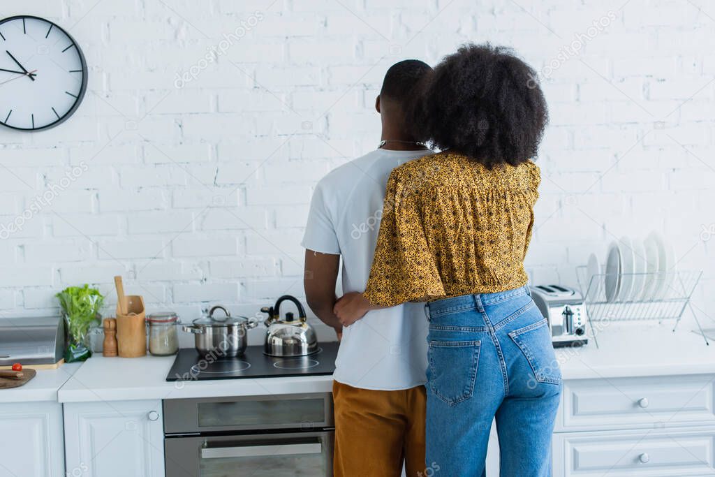 Back view of african american woman hugging man near stove in kitchen 