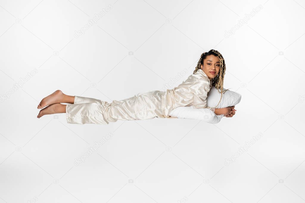 african american woman in silk pajamas levitating with pillow isolated on white