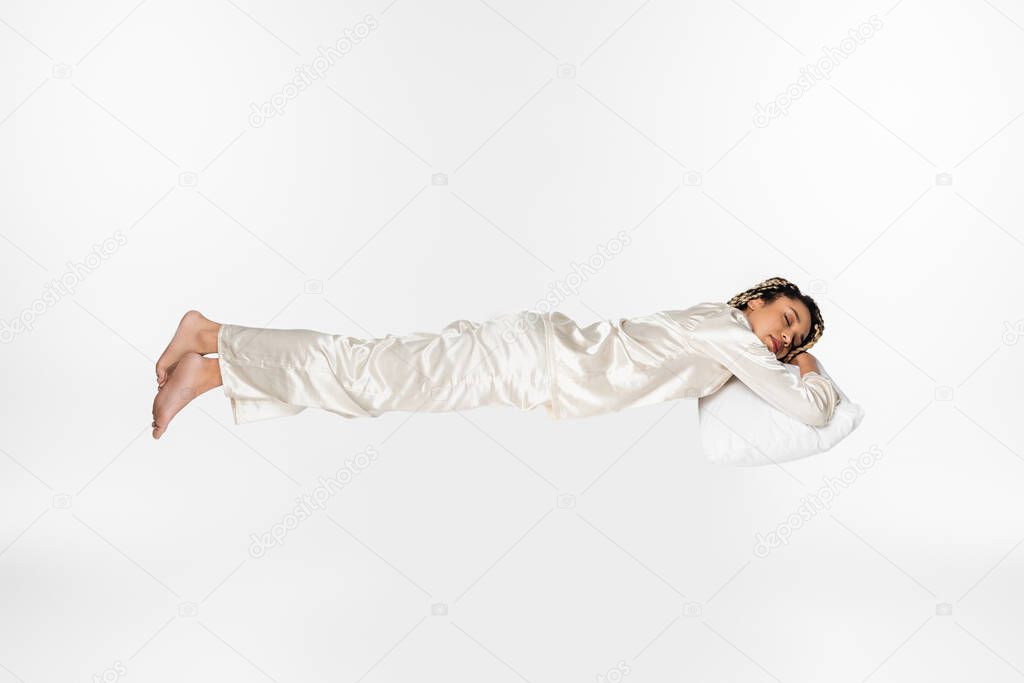 full length view of barefoot african american woman in silk pajamas sleeping while levitating isolated on white