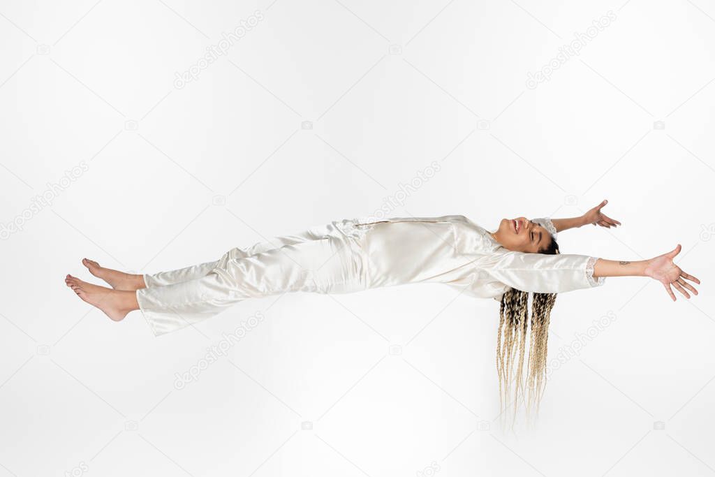 happy african american woman in silk pajamas sleeping while levitating in air isolated on white