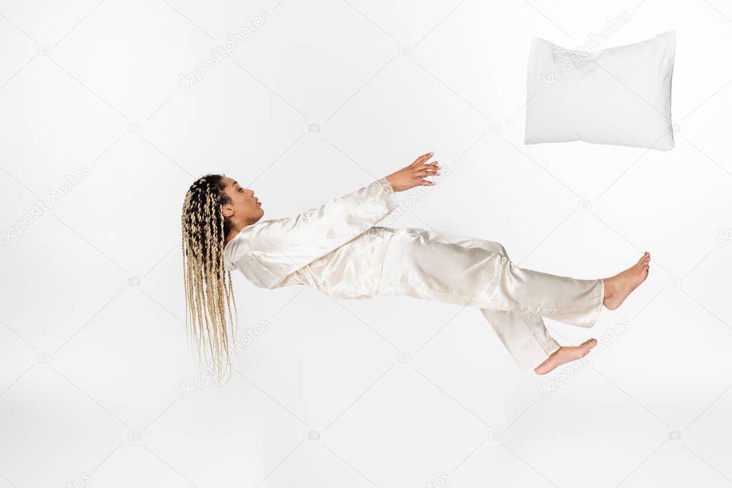 pillow flying away from african american woman levitating in silk pajamas isolated on white