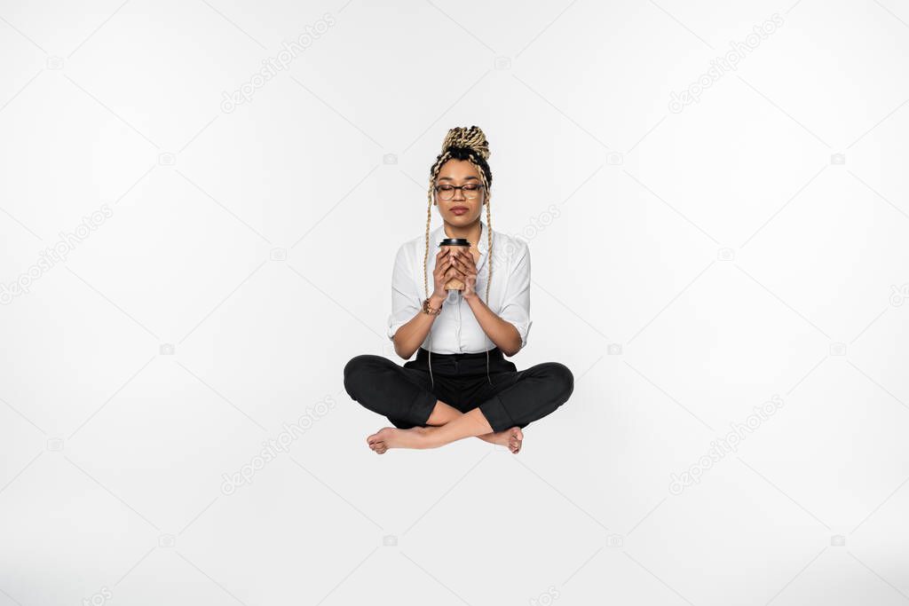 african american businesswoman holding coffee to go while levitating in lotus pose isolated on white