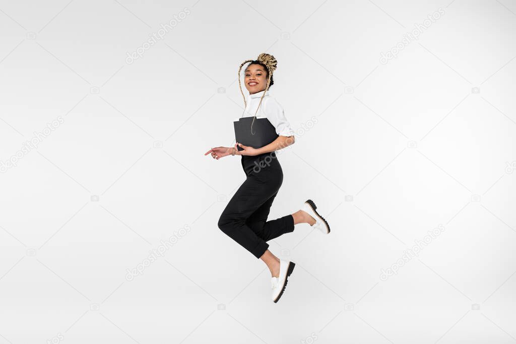 cheerful african american businesswoman looking at camera while levitating with paper folder isolated on white