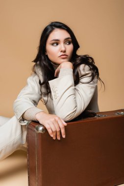 Takım elbiseli genç kadın bej rengi bavulun yanında poz veriyor. 