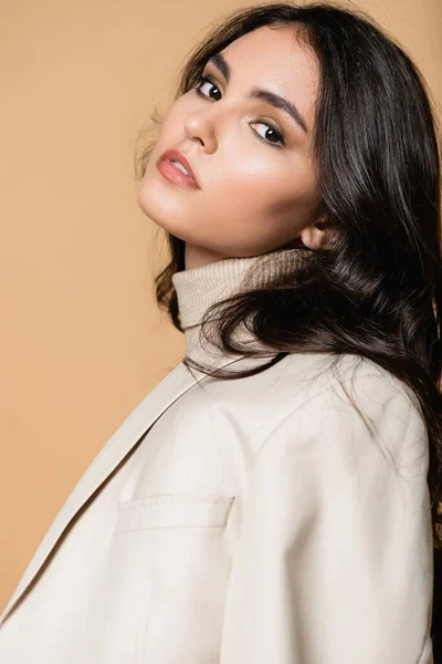 pretty model in turtleneck and blazer looking at camera isolated on beige