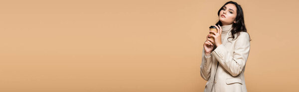 молодая красивая женщина в водолазке и пиджаке держа бумажный стаканчик изолирован на бежевый, баннер
