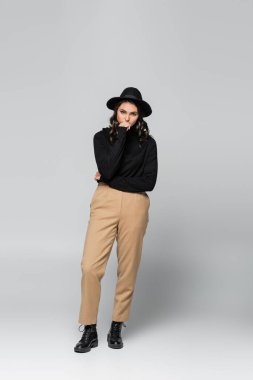 Uzun boylu, siyah fötr şapkalı, şık giyimli, ağzı kaplayan ve gri üzerinde poz veren genç bir model.