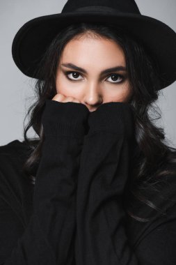 Siyah fötr şapkalı ve balıkçı yakalı genç manken kameraya bakarken gri boğazlı ağzını kapatıyor.