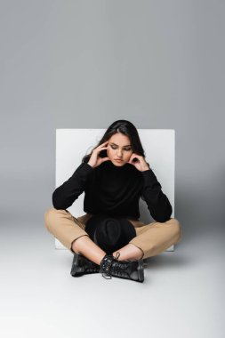 Siyah balıkçı yaka kazaklı şık bir model fötr şapkalı, gri küpün yanında oturuyor. 