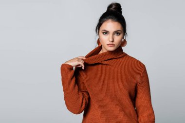 Genç bir kadın örgü kazağını düzeltiyor ve gri renkli kameraya bakıyor. 