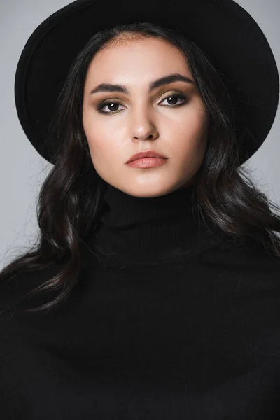pretty model in black fedora hat and turtleneck looking at camera isolated on grey