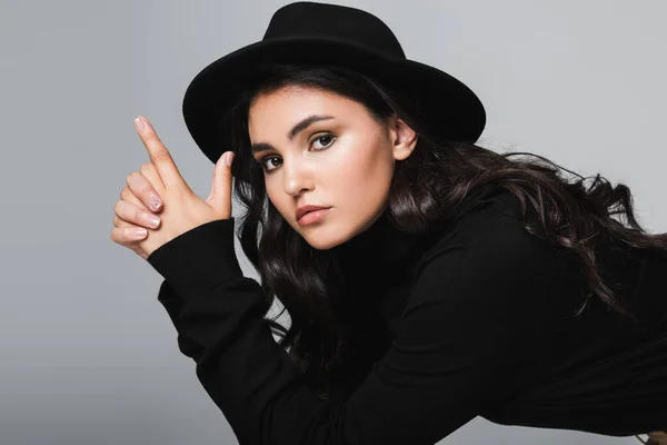 brunette and young model in fedora hat looking at camera while showing finger gun gesture isolated on grey