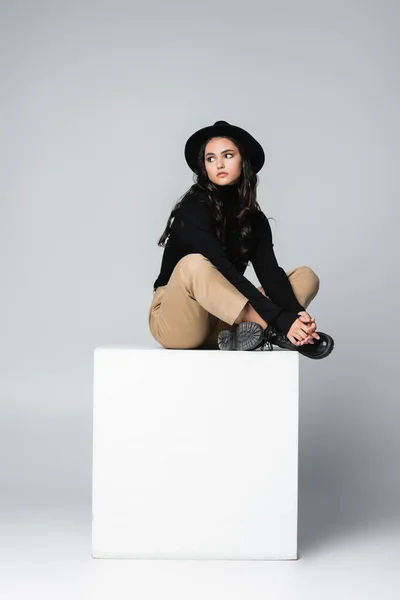 full length of stylish model in fedora hat sitting with crossed legs on white cube on grey