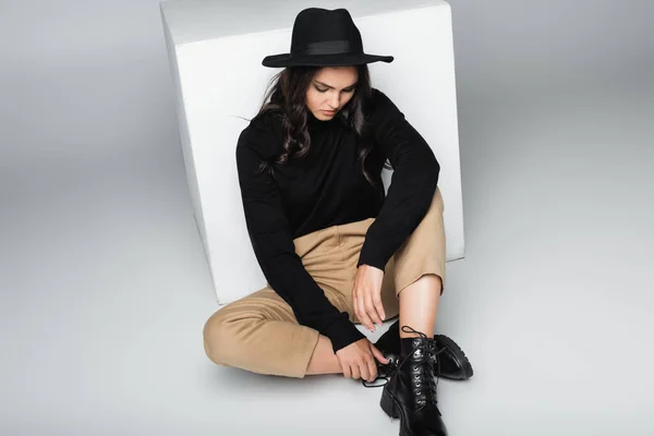 グレーの白い立方体の近くに座っているフェドーラの帽子の若いモデルの完全な長さ — ストック写真