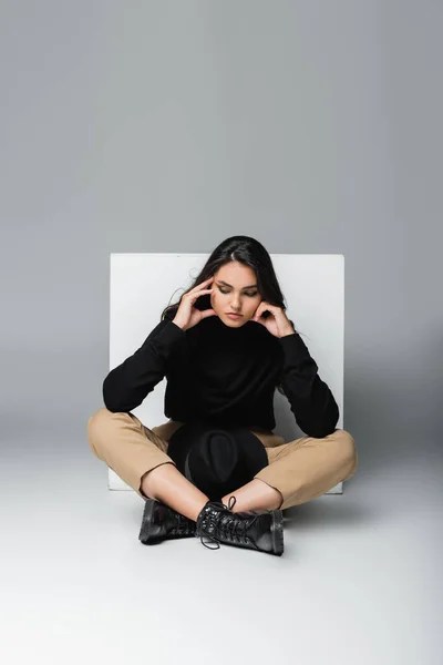 stylish model in black turtleneck with fedora hat sitting near white cube on grey