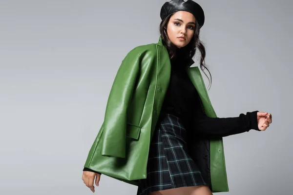 stylish woman in green leather jacket, checkered skirt and beret posing isolated on grey
