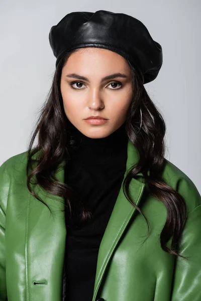 young model in black turtleneck, green jacket and beret looking at camera isolated on grey