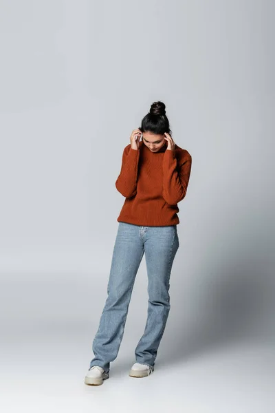 full length of young woman in sweater and denim jeans looking down on grey