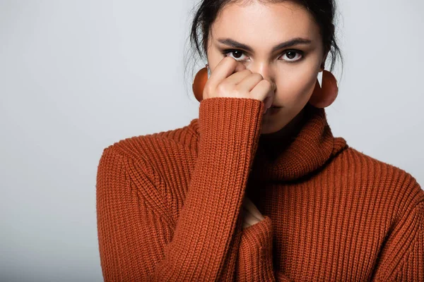 young woman in warm sweater looking at camera while covering face isolated on grey