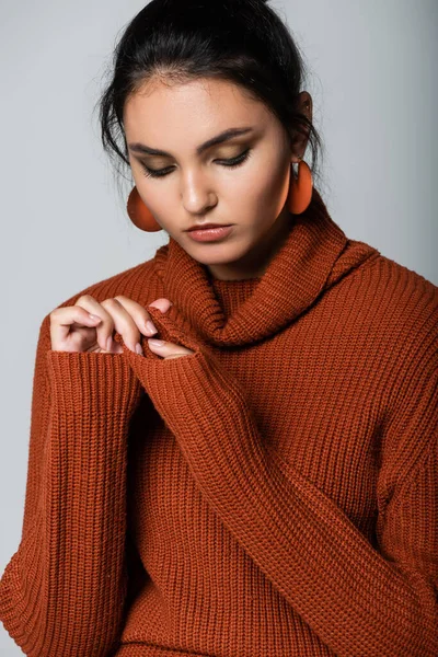 young woman in earrings and knitted sweater looking down isolated on grey