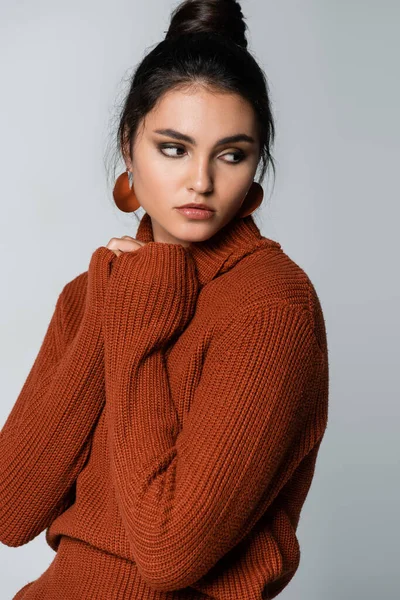 young woman in earrings and brown sweater looking away isolated on grey