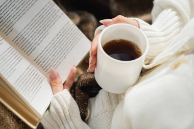 Elinde bir fincan sıcak çay tutarken kitap okuyan kadın manzarası.