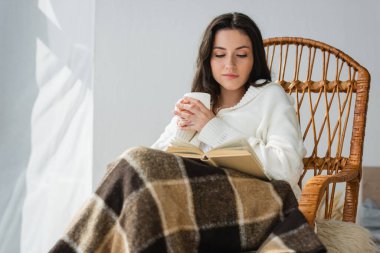 Esmer kadın hasır sandalyede oturmuş elinde çay ve kitap okuyor.