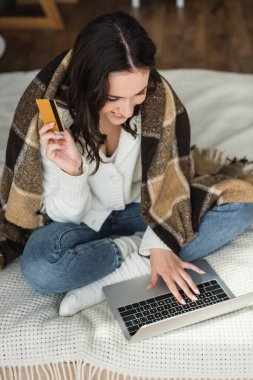 Kredi kartı tutan ve laptopta kareli battaniyenin altında yazan mutlu bir kadın.