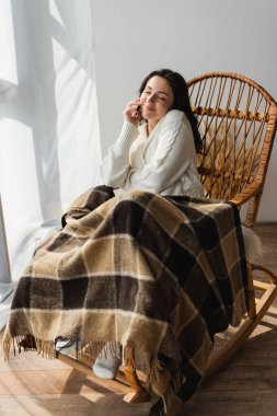 Sıcak battaniyenin altında sallanan sandalyede oturan ve akıllı telefondan gözleri kapalı konuşan memnun bir kadın.