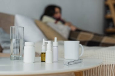 selective focus of containers with medication, thermometer and drinks on bedside table near sick woman on blurred background clipart