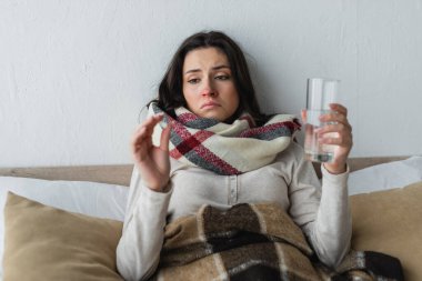 sick and sad woman holding glass of water and pill while lying under plaid blanket clipart