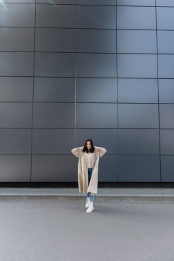 Beyaz çizmeli ve bej renkli yağmurluklu gülümseyen kadın ellerini başının arkasına koyup yüksek duvarın yanında poz veriyor.