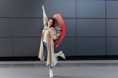 Kırmızı şemsiyeli bej yağmurluklu neşeli kadın elini kaldırdı. 