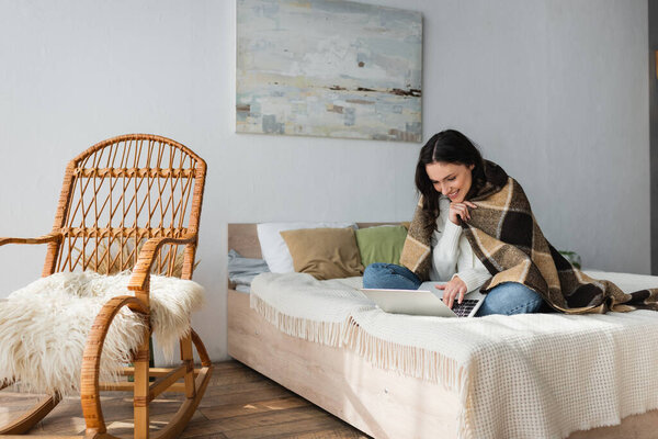 cheerful woman using laptop on bed near wicker chair 