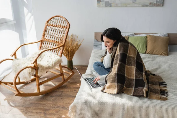 Lächelnde Frau Sitzt Auf Bett Neben Schaukelstuhl Und Benutzt Laptop — Stockfoto