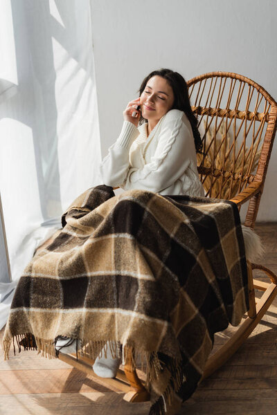 довольная женщина сидит в кресле-качалке под теплым одеялом и разговаривает на смартфоне с закрытыми глазами