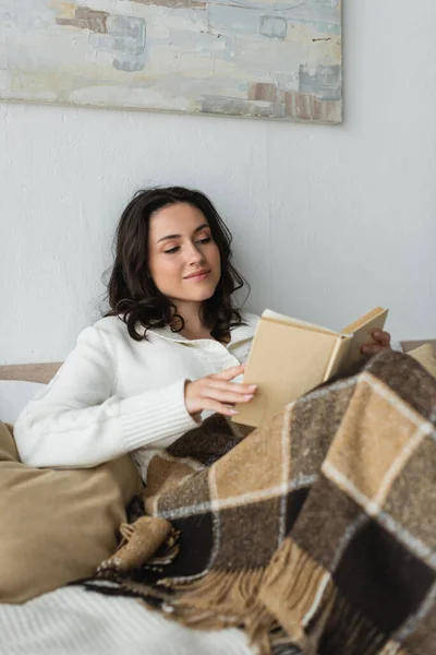 穿着白色羊毛衫躺在温暖的毯子下躺在床上看书的积极的年轻女人 — 图库照片